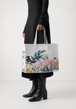 Persona con vestito nero e stivali che tiene una grande borsa a spalla bianca con stampa floreale colorata e manici corti.