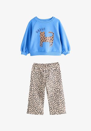 Mėlynas džemperis su rudu dėmėtu katinu ir užrašu „HELLO“, derinamas su smėlio spalvos kelnėmis, kuriose yra juodų nereguliarių dėmių.