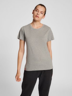 Kvinde med lys hud og håret bundet tilbage iført en enkel grå t-shirt med korte ærmer og sorte bukser, stående mod en ensfarvet baggrund.