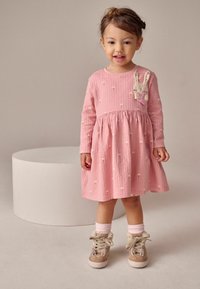 Růžové šaty s texturovanou tkaninou, dlouhými rukávy a nařaseným pasem. Akcentované malými vzory a detailem králíčka na hrudi. Hnědé boty s králičími ušima.