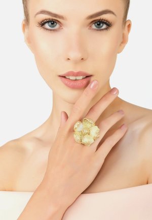 Gold floral ring featuring textured petals and a central embellishment, worn on a finger against a neutral background.