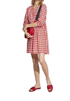 Frau trägt ein rot-weiß gemustertes Kleid, rote Pantoletten mit Schnallen und eine rote Umhängetasche mit schwarzem Riemen.