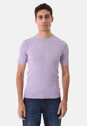 Uomo con capelli corti e scuri, indossa un maglione aderente a maniche corte color lavanda a coste e jeans blu scuro, in piedi davanti a uno sfondo semplice.