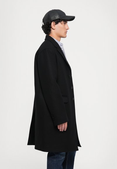 Black wool overcoat with a tailored fit, notched lapels, and five button closures. Paired with a black leather cap and blue jeans.