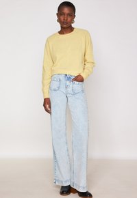 Donna in piedi, che indossa un maglione giallo chiaro, jeans a gamba larga blu chiaro con tasche frontali, stivaletti neri con tacco e orecchini a cerchio dorati.