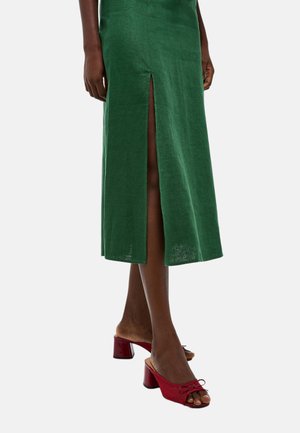 Jambes portant une jupe midi verte avec une fente devant et des mules rouges à talons blocs ouverts, sur un fond clair uni.