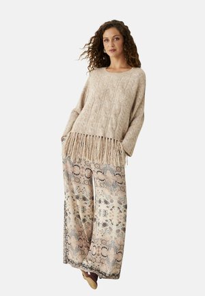 Mujer con cabello rizado que lleva un suéter beige de punto con flecos y un pantalón ancho con estampado floral, de pie con las manos en los bolsillos.