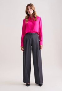 Fuchsia blouse met knopen en kraag, gecombineerd met grijze wijde broek met strepen. Slim fit met strakke lijnen, gedragen met zwarte hakken.