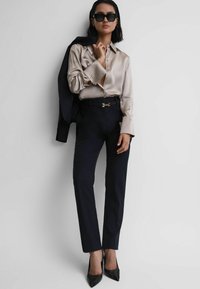 Blusa beige in seta con bottoni, pantaloni neri su misura e décolleté neri con punta. Blazer nero appoggiato sulla spalla, occhiali da sole neri oversize.