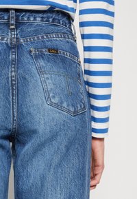 Modré džínové kalhoty s vybledlým povrchem, vysokým pasem a zadní kapsou s malou značkovou etiketou. Rukáv pruhované košile je částečně vidět.