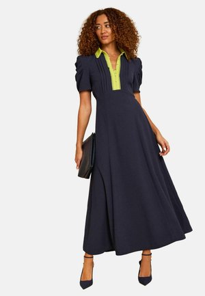 Frau trägt ein marineblaues Midikleid mit Puffärmeln und limonengrünem Kragen, hält eine schwarze Clutch und steht vor einem einfarbigen Hintergrund.