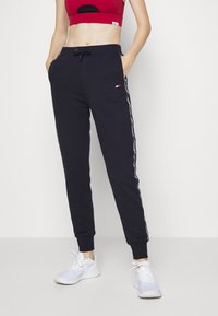Marineblaue Sweatpants mit elastischen Bündchen, Kordelzugbund, seitlichen Taschen und kontrastierenden vertikal Streifen. Kombiniert mit weißen Sportschuhen.