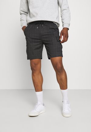 Mann trägt graue karierte Shorts, weiße Turnschuhe und weiße Socken, steht mit teilweise in den Taschen platzierten Händen vor einem einfarbigen Hintergrund.