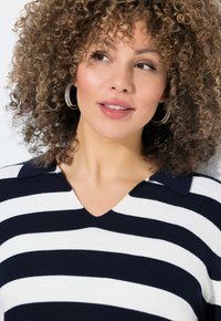 Marinblå och vit randig polo med V-hals och krage, med ett texturerat stickat mönster. Modellen har lockigt hår och hoopörhängen.