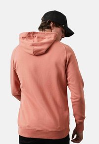 Sudadera de color coral con cordón, puños y cintura acanalados. Presenta una textura suave y un bolsillo tipo canguro, usada con una gorra negra.