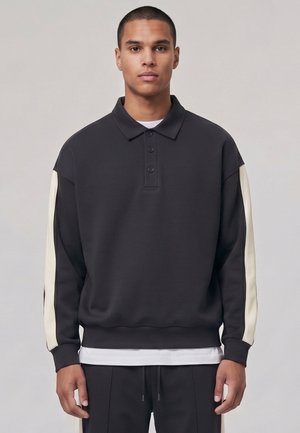 Jeune homme aux cheveux courts portant un polo noir à manches longues avec des rayures blanches sur les manches et un pantalon noir assorti.