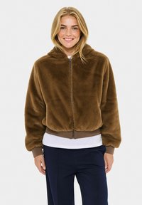 Hnědá fleecová bunda s kapucí, se zipem a žebrovaným lemem. Měkká textura a volný střih. Nosí se s bílou košilí a tmavými kalhotami.