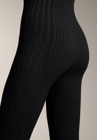 Približeni prikaz crnih rebrastih pletenih tajica s istaknutim teksturiranim dijagonalnim uzorkom na bedru i bokovima na neutralnoj pozadini.