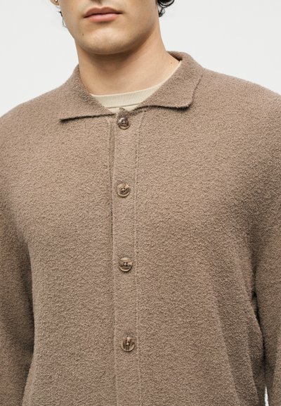 Άνδρας που φοράει μπεζ υφασμάτινη ζακέτα με κουμπιά πάνω από ένα ανοιχτό μπεζ μπλουζάκι με στρογγυλή λαιμόκοψη, δείχνοντας λεπτομέρειες για το γιακά και το πάνω μέρος του σώματος.