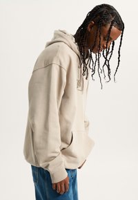Felpa beige con tasca frontale e cappuccio con cordino, abbinata a jeans in denim blu. Il tessuto appare morbido con una vestibilità rilassata.