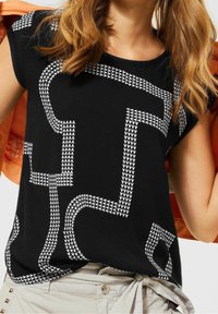 Haut noir à manches courtes avec un motif géométrique blanc ressemblant à des lignes et des triangles interconnectés. Le tissu semble doux et lisse.