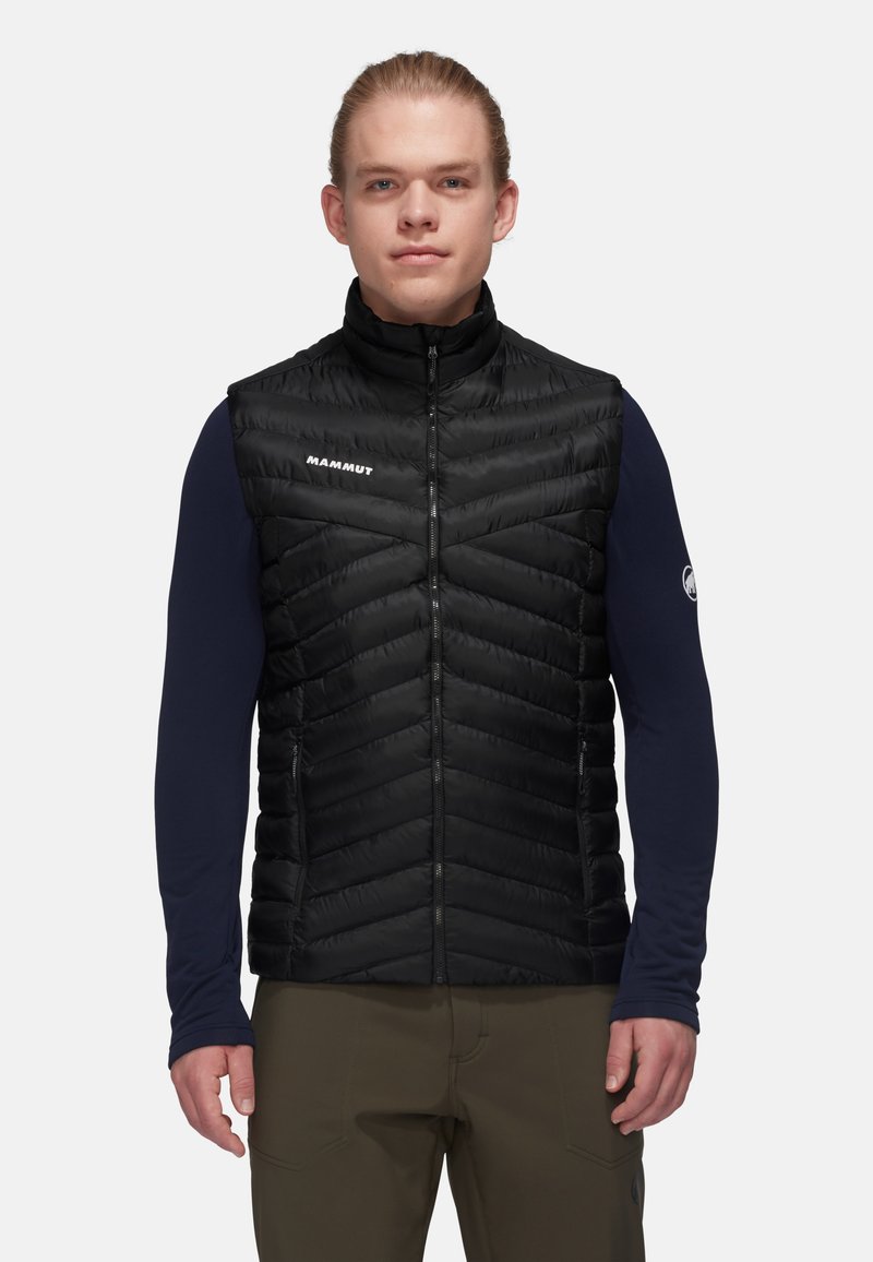 Schwarze isolierte Weste mit Zickzackmuster, Reißverschluss und hohem Kragen. Über einem langärmeligen Marineblauen Hemd getragen. Minimalsitisches Branding sichtbar.