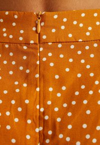 Oranžová látka s bílými puntíky, s uzávěrem na zip. Materiál má hladkou texturu, která zdůrazňuje hravý vzor.