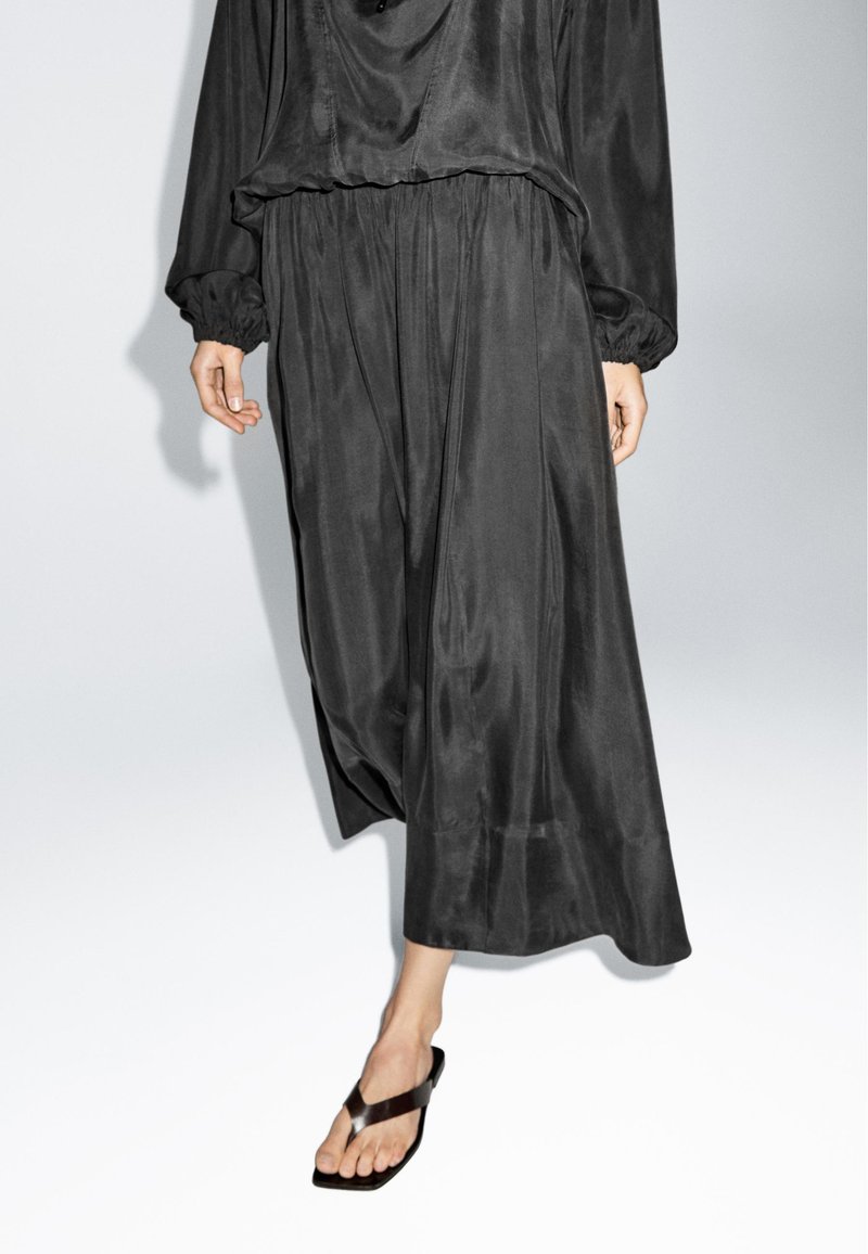 Vrouw draagt losse zwarte satijnen broek, bijpassende top met lange mouwen en zwarte teenslippers tegen een effen lichte achtergrond.