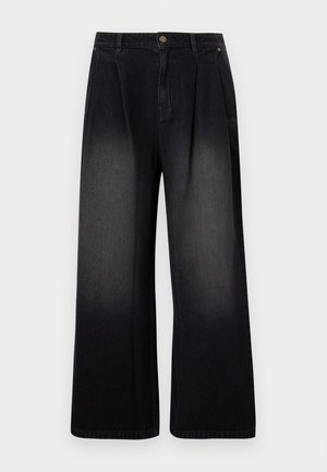 Uomo che indossa jeans neri sbiaditi a gamba larga e sneakers bianche, in piedi con le braccia rilassate lungo i fianchi davanti a uno sfondo neutro.