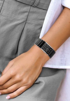 Schwarze rechteckige Uhr mit silberner Hardware, ausgestattet mit einem strukturierten Armband und einem minimalistischen schwarzen Ziffernblatt mit silbernen Stundenmarkierungen.