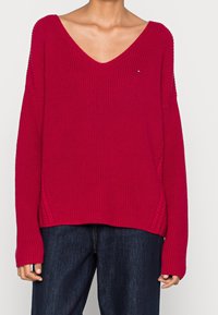 Rode gebreide trui met een diepe V-hals, losvallend model en geribbelde textuur. Heeft zijslits en lange mouwen, gecombineerd met een donkere spijkerbroek.