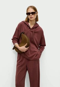 Donna che indossa una felpa maroon e pantaloni, mentre tiene una borsa a forma di mezzaluna marrone e indossa occhiali da sole rettangolari scuri.