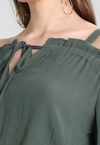 Zelená, lehká blůza s designem bez ramene, nastavitelnými detaily na uvázání a texturovanou tkaninou. Obsahuje široké ramínka a volný střih.
