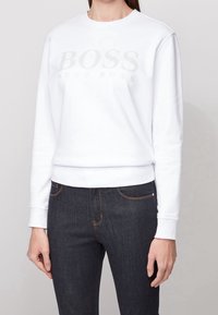 Sweatshirt blanc à col rond, avec un logo gris subtil "BOSS HUGO BOSS" sur la poitrine, porté avec un jean en denim foncé.