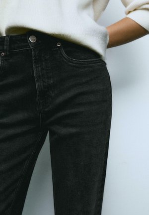 Jean en denim noir avec une coupe slim, présentant un bouton métallique et des coutures visibles. Porté avec un pull en tricot de couleur claire.