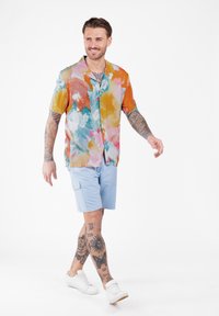 Mann läuft und trägt ein mehrfarbiges kurzärmliges Hemd, hellblaue Shorts, weiße Sneaker sowie sichtbare Tattoos an Armen und Beinen.