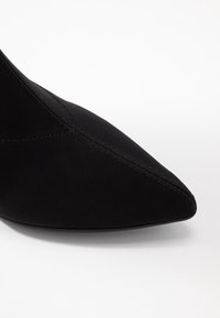 Chaussure noire à bout pointu en matériau doux, avec des accents cousus et une silhouette élégante, sur fond blanc.