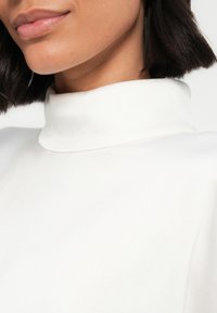 Valkoinen korkea kaulus, joka esittelee sileän tekstuurin. Lähikuva kaula-aukosta ja ihosta, korostaen puhdasta, minimalistista muotoilua.