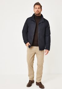 Marineblaue gepolsterte Jacke mit Reißverschluss, einer Brusttasche und einem Kragen, getragen über einem braunen Rollkragenpullover. Beigefarbene Chinos und braune Schnürschuhe vervollständigen das Outfit.