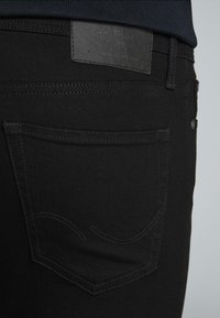 Jeans de denim negro con un parche de cuero en la parte trasera de la cintura, bolsillos estándar, detalles de costura sutiles y una textura suave.