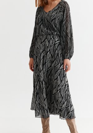 Musta pitkä mekko, jossa valkoinen lehtikuvio, pitkät läpikuultavat hihat, V-kaula-aukko, vyötärö leikattu ja liehuva helma.