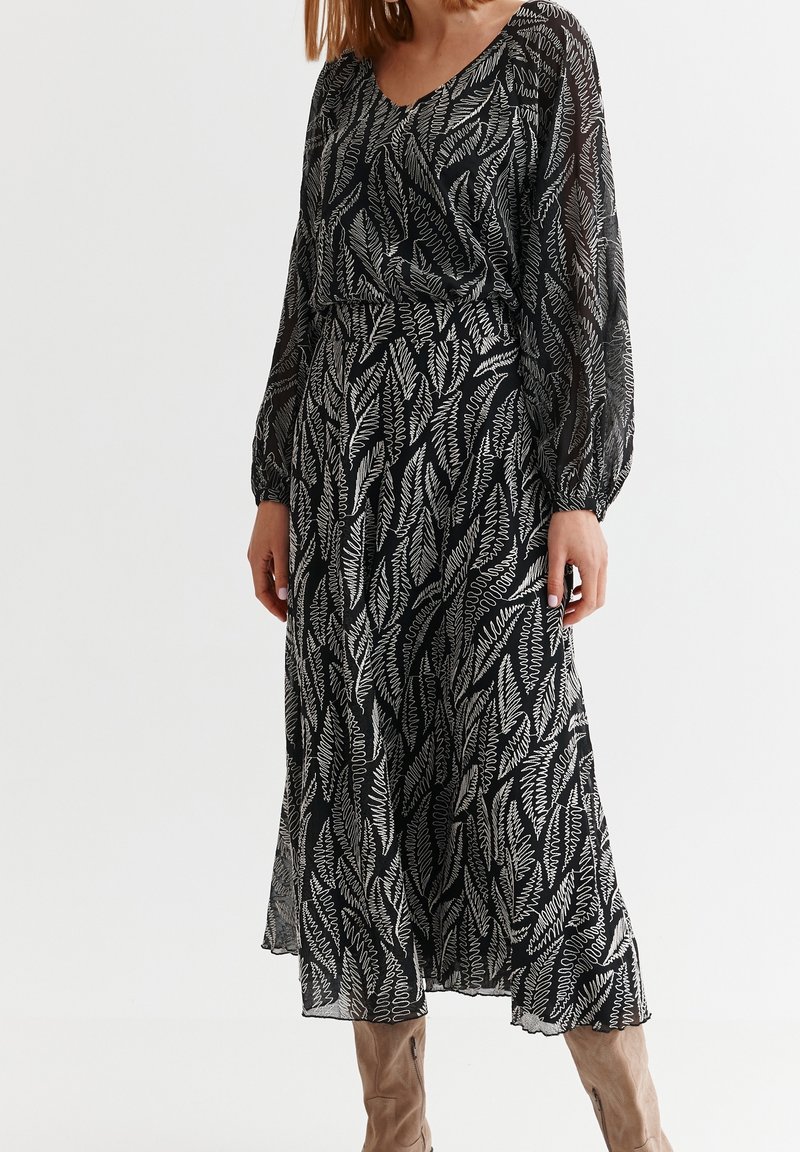 Robe longue noire avec motif de feuilles blanches, manches longues transparentes, décolleté en V, taille cintrée et bas fluide.