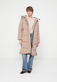 Persona con un abrigo acolchado largo beige sobre un suéter crema y jeans azules, de pie contra un fondo blanco liso.