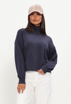 Vrouw met een beige pet, een marineblauwe coltrui en witte broek, staand met één hand bij haar taille en de andere ontspannen.