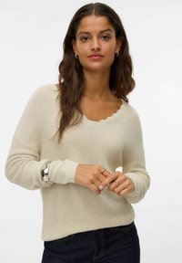 Maglione beige lavorato a maglia con scollatura a V e bordo ondulato, maniche lunghe e polsini a coste, abbinato a jeans in denim scuro.