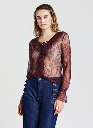 Blusa in pizzo borgogna con scollatura arricciata, maniche lunghe e dettaglio a fiocco. Abbinata a pantaloni in denim blu a vita alta con bottoni dorati.