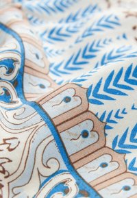 Zoom su un tessuto con motivi di foglie blu e un bordo decorativo beige con vortici e piccoli punti blu su sfondo bianco.