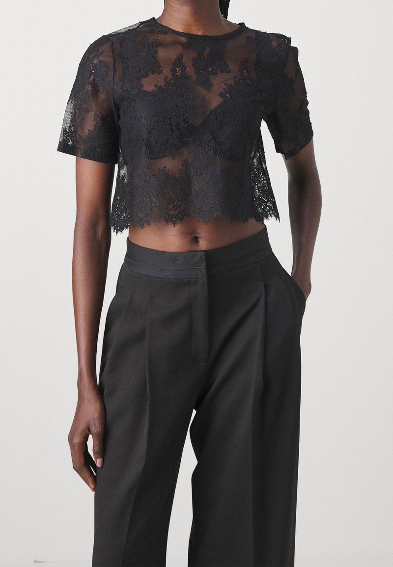 Crop top court en dentelle noire à manches courtes porté avec un pantalon noir plissé taille haute, une main dans la poche sur un fond clair uni.