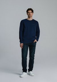 Granatowy sweter z okrągłym dekoltem, wykonany z miękkiego materiału, noszony z ciemnymi dżinsami i białymi trampkami. Minimalistyczny design z subtelnymi detalami.