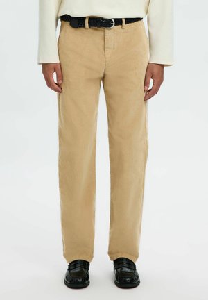 Pantalon en velours côtelé beige porté avec une ceinture tressée noire et des mocassins noirs texturés, associé à un haut crème à manches longues.